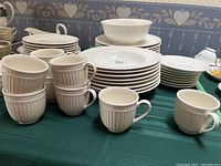 Photo showing stacked white porcelain plates and bowls featuring ribbed vertical patterns along edges, along with coffee mugs with handles and ribbed texture.