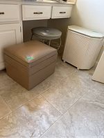 Photo showing vanity stool with beige faux leather top and storage feature, laundry hamper in woven pattern material, and faux leather ottoman in beige/tan color grouped under a cabinet.