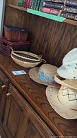 Photo showing four hats and four baskets arranged on a wooden cabinet. Hats include white canvas tilly hat with chin strap, straw hat with blue floral band, two woven wide-brimmed hats with patterns. Baskets include oval, round woven, decorative bead basket, and rectangular reddish basket.