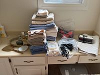 Photo showing stacked assortment of bath towels and bath mat, plastic bags with hair dryers and curling iron, and GE alarm clock with manual.