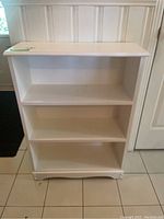 Front full view of white wood bookshelf with three shelves showing intact structure but surface wear.