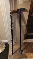 Two walking canes standing upright; left cane is dark handmade wood with branch-like texture, right cane is blue metal adjustable with black handle and three-point rubber tip.