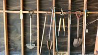Full view of garden hoe, shovel with black plastic grip, garden fork, and various rakes leaning against the wall.
