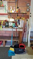 View of garage area with cleaning tools including Vileda mop bucket, mop, dustpan, broom, shelf with spray bottles, cans, and hardware containers