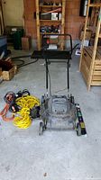 Photo of used black Jobmate electric lawn mower with electrical cables coiled beside it, displayed in a garage.