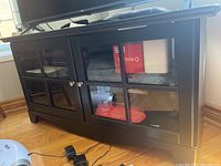 Front angled view of the black media cabinet showing two glass doors with grid pattern and metal knobs, resting on wooden floor.