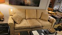 Beige microfiber three seat sofa with two matching throw pillows, shown from front angle with lamp and art in background.