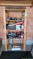 Front view of pine shelving unit against brick wall showing items on four shelves including tools, plumbing parts, spray paint cans, and boxes.