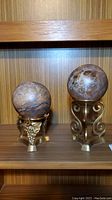 Two stone spheres mounted on decorative brass holders displayed on a wood shelf, showing size difference and material details.