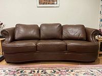 Front view of a brown leather 3-seater sofa with three seat cushions, curved front, and rolled arms, placed on a floral rug in a living room.