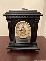 Front view of black wooden mantel clock showing classical design, clock face with Roman numerals, and visible gold-colored gears behind a glass panel.
