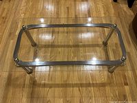 Photo showing a rectangular glass top coffee table with four shiny metal cylindrical legs and metal frame support on a wooden floor.