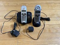 Two Siemens cordless phone handsets with numeric keypads and small displays on a wooden floor, along with their black base station and two power adapters with cords.