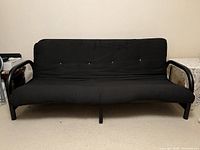 Black futon bed in sofa position with black fabric and metal frame with wooden armrests.
