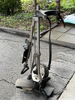 Folded golf pull cart leaning against an outdoor sidewalk, showing metal frame, wheels and strap in clear view.