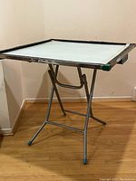 Foldable mahjong table with white play surface, metal folding legs, and dark frame with green corner protectors
