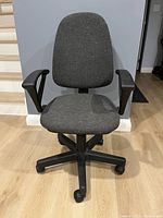 Front view of gray fabric office chair with black armrests and caster wheels, placed on wood floor.