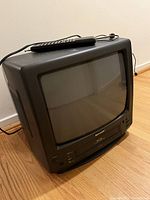 Black Sharp CRT TV with integrated VHS player positioned on a wooden floor against a white wall. Remote control is placed on top of the TV unit.