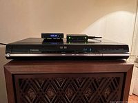 Photo of a black Toshiba DVD player placed on a wooden stand with two small electronic devices on top that are not included in the sale.