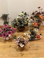 Photo showing six different artificial flower arrangements in varying colors and container styles on a wooden floor.