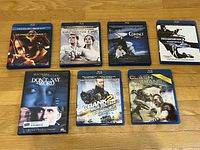 Photo shows 7 Blu-Ray movie cases laid out on a wooden surface with visible titles and cover art.