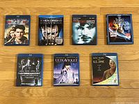 Seven Blu-Ray movie discs arranged on wooden floor showing front covers for Top Gun, X-Men Origins Wolverine, The Butterfly Effect, The Last Airbender, Daybreakers, Ultraviolet, and G.I. Jane.