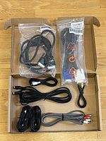 Top-down photo of a box containing various cables, some in plastic packaging, showing USB cable, power cables, and RCA audio cables.