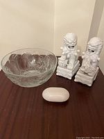Clear glass bowl with floral pattern, two white porcelain lion sculptures on decorative bases, soap-shaped glass weight.