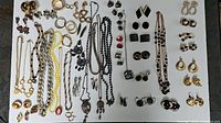 Top-down image showing all necklaces and earrings laid out on flat surface in rows. Gold-tone, black, and mixed metal-tone jewelry pieces are visible including chains, beads, and various earring styles.