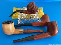 Four smoking pipes resting on a clear glass ashtray with pipe cleaners in background