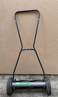 Photo of the full push mower showing black metal frame and wheels in front of a beige wall.
