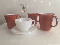 Three red-orange Pyrex mugs, white FireKing cup and saucer, vintage glass flute with cherry design shown on table.