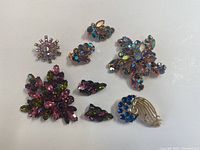 Lot of nine rhinestone pins and clip-on earrings, various colors and shapes displayed on a white surface.