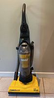 Full view of the upright vacuum showing yellow base and clear dust container with hose attached.