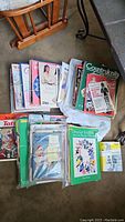 Top down view of a pile of vintage crafting and painting books and magazines on the floor beside a wooden chair, showing colorful spines and covers.