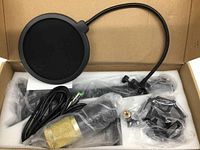 Microphone kit in cardboard box showing gold condenser microphone, pop filter, shock mount, microphone stand arm and XLR cable in plastic wrapping.