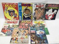 Photo of seven Mad Magazines and two Cracked Magazines, arranged standing upright, covers visible showing various themes including TV face, Ghostbusters parody, wrestling characters, and group illustrations.