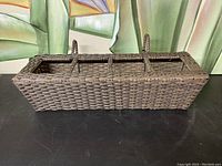 Rectangular wicker balcony box with three compartments and a central handle on black surface with green leaf background.