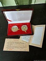 Two silver coins/medals in clear protective capsules with yellow ribbons, placed inside a red velvet-lined wooden display case, accompanied by informational documents.
