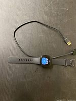Full view of Fitbit Versa 3 smartwatch with black adjustable silicone band and attached USB charging cable laid on a dark surface.
