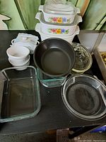 Vintage Pyrex bakeware including 3 nesting casseroles with floral design, clear pie dish, rectangular baking dish, black pot, and white and brown tea cups.
