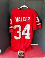Back view of red jersey on white hanger against black backdrop showing 'WALKER' name and large white number 34 with signature on 4; small printed photo at base.