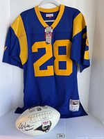 Front view of royal blue and yellow St. Louis Rams jersey #28 by Mitchell & Ness with tags and Wilson football at base
