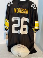Back view of black Pittsburgh Steelers jersey #26 with 'WOODSON' nameplate, signed by Rod Woodson on the numbers; autographed white panel football positioned below.