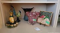 Full shelf view showing the lot with glass rooster figurine, decanter set with glasses, two art glass pieces (blue vase and pink bowl), and decorative tile on small stand.