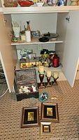 Open cabinet shelf with various decorative items including candles, candle holders, framed pictures, faux flowers in pots, a decorative box, and figurines