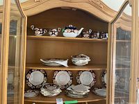 China set displayed in wooden cabinet across three shelves: top shelf holds teapot, sugar bowl, creamer and cups; middle and lower shelves hold stacks of plates and saucers.