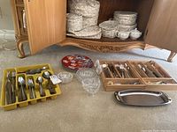Photo showing the full assortment of cutlery in trays and wooden box, two glass candy dishes, salt and pepper shakers, two decorative plates, and a steel serving tray placed on carpet in front of wooden cabinet