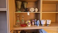 Two crystal decanters, three porcelain china flower arrangements with chips, red decorative art glass ashtray, two Pottery Barn urns with peeling glaze and other decorative vases shown on a wooden shelf.