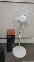 Full view of Honeywell white pedestal fan standing on round base next to Sunbeam tower heater fan and box behind.